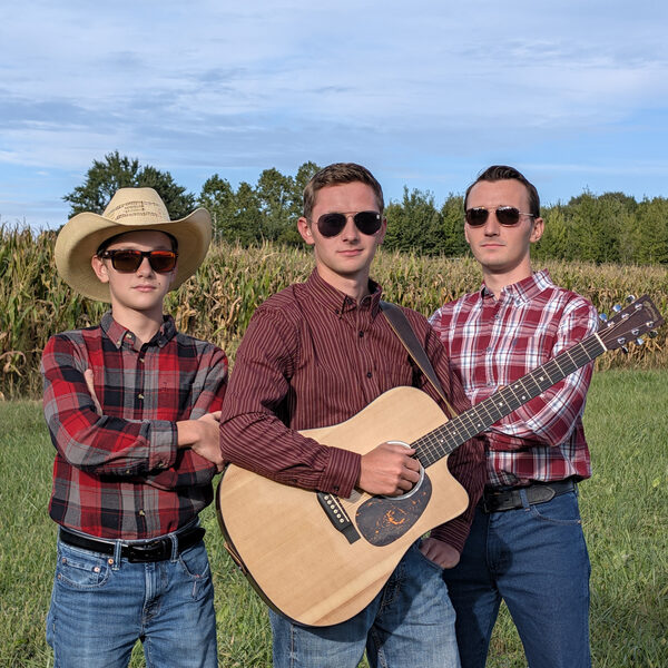 The three Stier Brothers in plaid shirts, one holding an acoustic guitar, in front of a cornfield